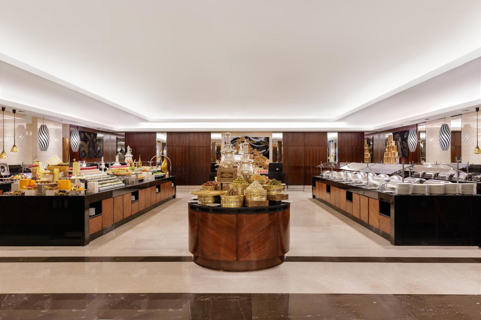 Swissotel Makkah Mecca Exterior photo The photo shows a spacious buffet dining area. The setting features elegant decor with polished surfaces and warm lighting. In the center, there is a large circular display of intricate food containers, likely showcasing an assortment of dishes. Surr