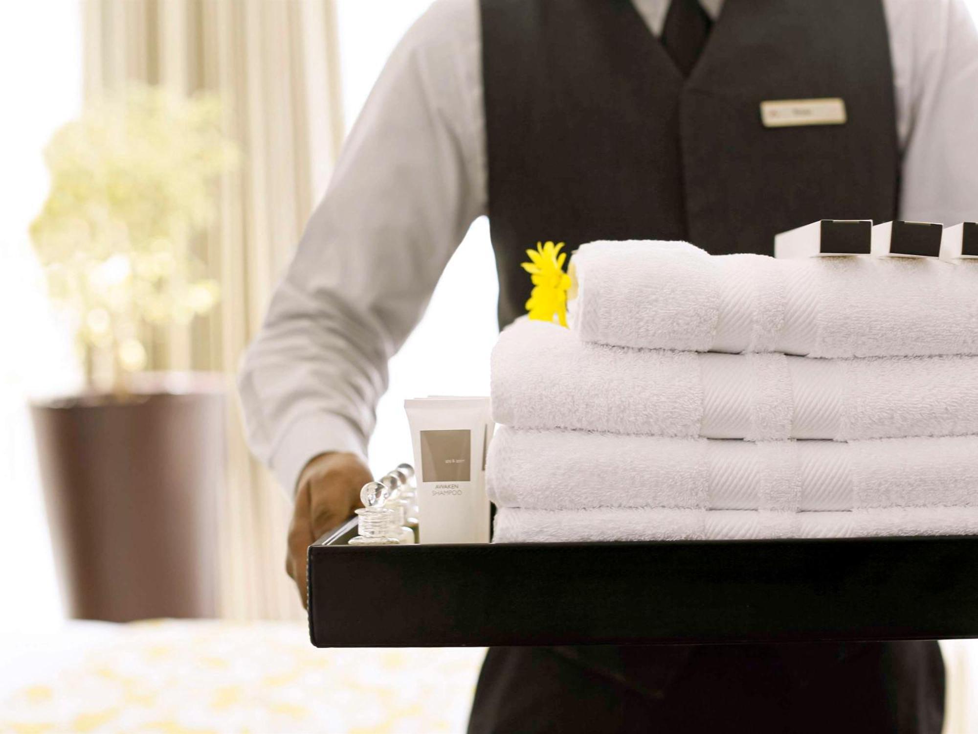 Swissotel Makkah Mecca Exterior photo The photo shows a person, likely a hotel staff member, holding a tray filled with neatly stacked white towels. The tray also contains a small tube of cream or lotion and several black and white square containers, possibly toiletries. A yellow flower 