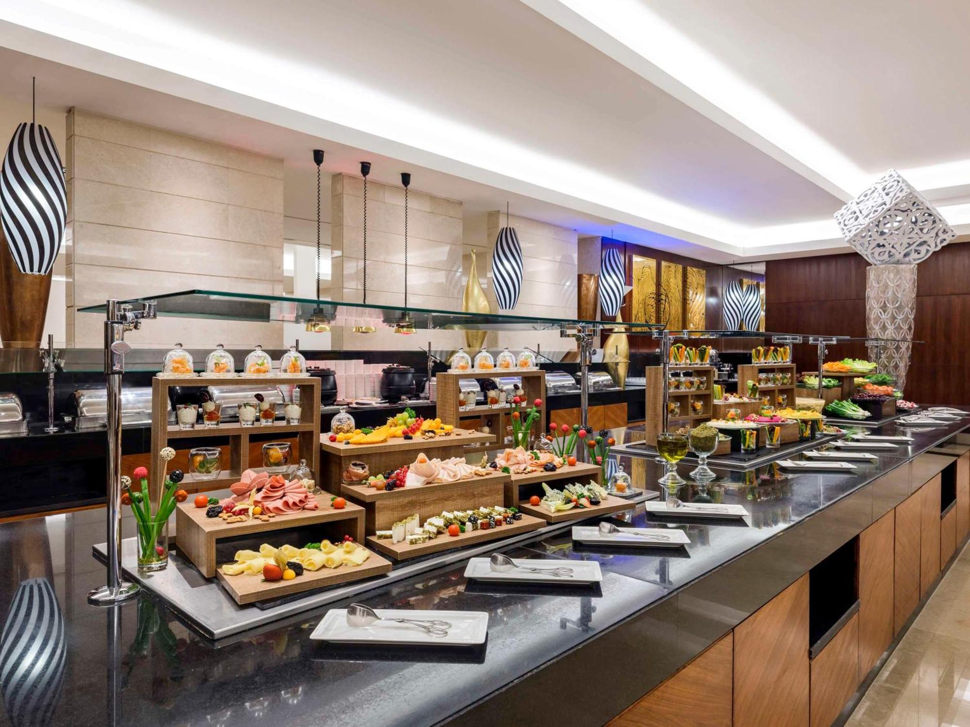 Swissotel Makkah Mecca Exterior photo The photo shows a buffet setup in a restaurant or hotel. There are several serving stations displaying a variety of foods. The setup includes tiered wooden racks filled with an assortment of dishes, likely featuring fruits, desserts, and appetizers. 