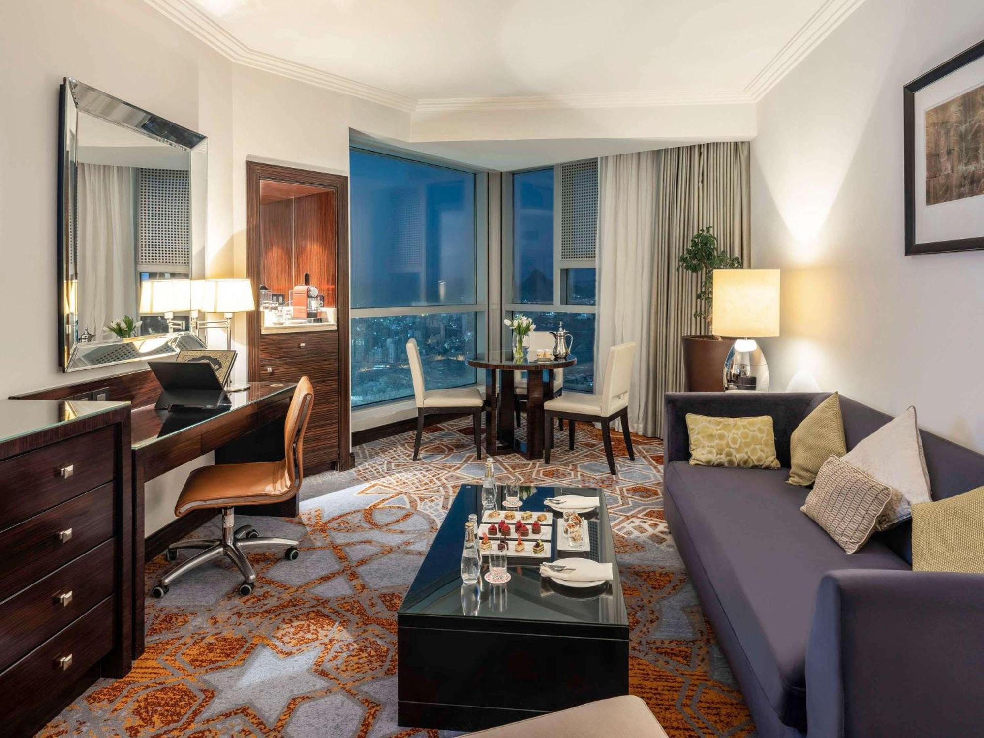 Swissotel Makkah Mecca Exterior photo The photo shows a well-furnished hotel suite or living area. It features a comfortable seating arrangement with a modern sofa and a coffee table. There is a dining area with a round table and chairs. On one side, there is a desk with a chair next to 